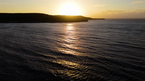 Panoramic-drone-ascend-as-sun-sets-behind-tropical-mounain-casting-light-ray-across-open-ocean