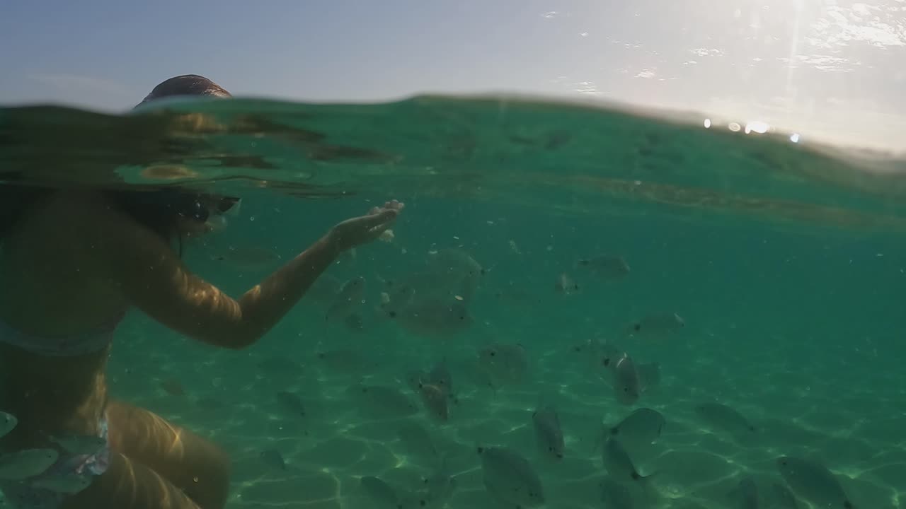 빵으로 먹이를 주는 물고기 떼에 둘러싸인 다이빙 마스크를 쓴 귀여운 빨간 머리 소녀의 수선 경치