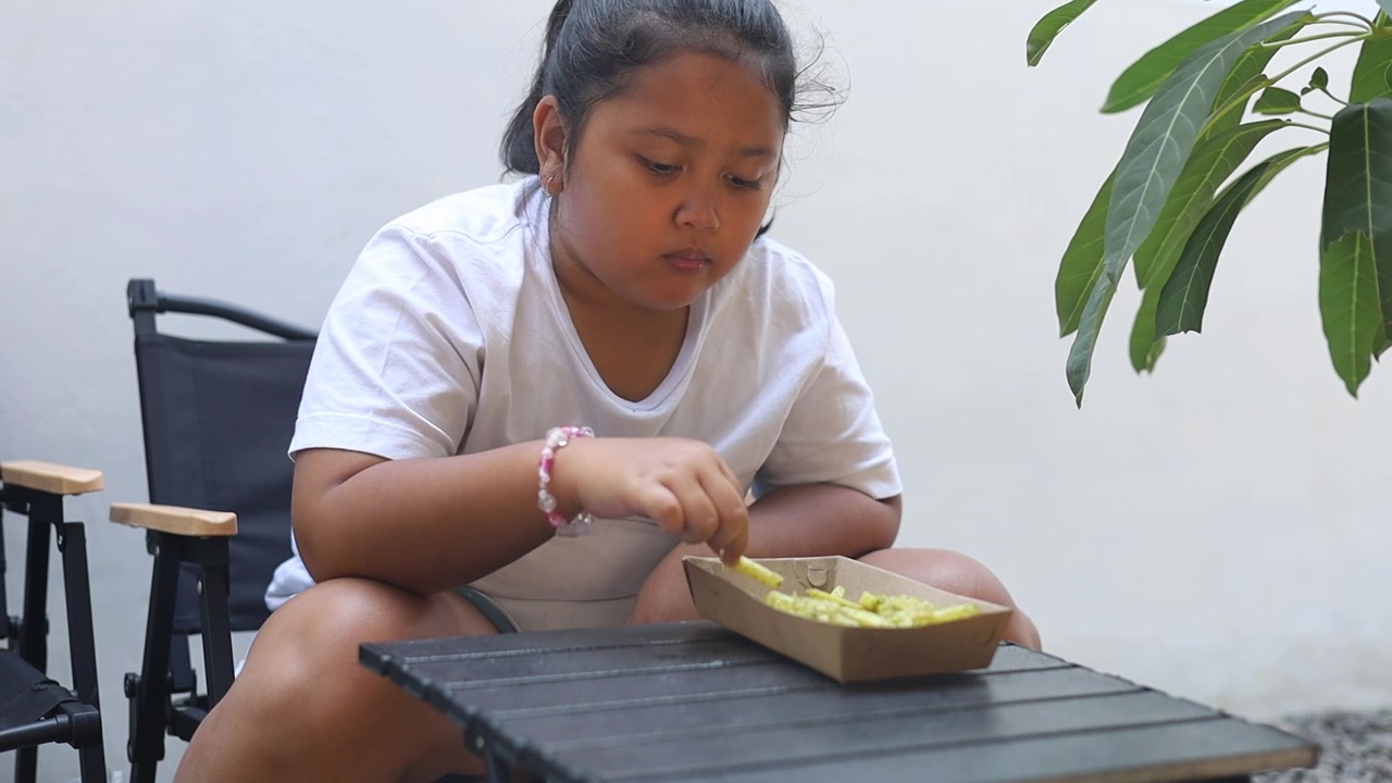 Ragazza asiatica che mangia patatine fritte mentre è seduta all'aperto a casa, catturando un momento casuale dell'infanzia, l'ora dello spuntino e lo stile di vita di una famiglia moderna.