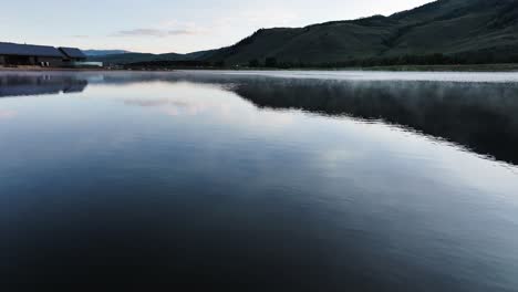 Temprano-En-La-Mañana-La-Condensación-De-La-Niebla-Se-Eleva-Desde-Un-Gran-Lago-En-Colorado-Con-Una-Casa-En-El-Lago-Y-Montañas-En-El-Fondo-Plataforma-Rodante-Aérea