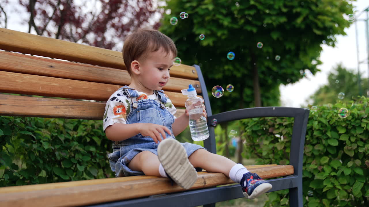 Cute Caucasian toddler sits on the bench holding a water bottle. Lovely boy is happy about soap bubbles flying around and tries to catch them.
