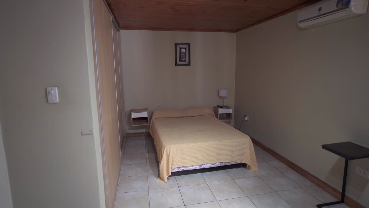 Simple house bedroom interior with double bed and wooden ceiling under soft lighting.