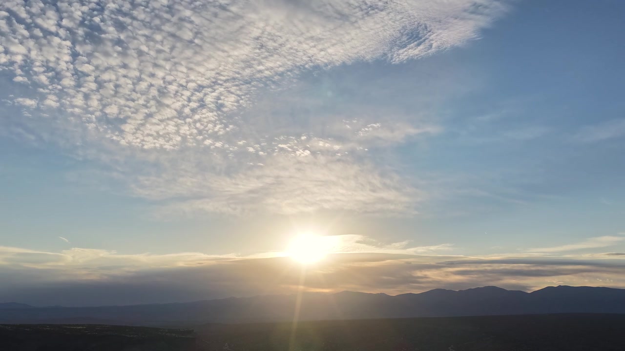 4K drone timelapse captures a sunrise bursting over a mountain cloud layer. Cinematic aerial motion reveals golden morning rays piercing through speckled clouds moving overhead across a vast vista.