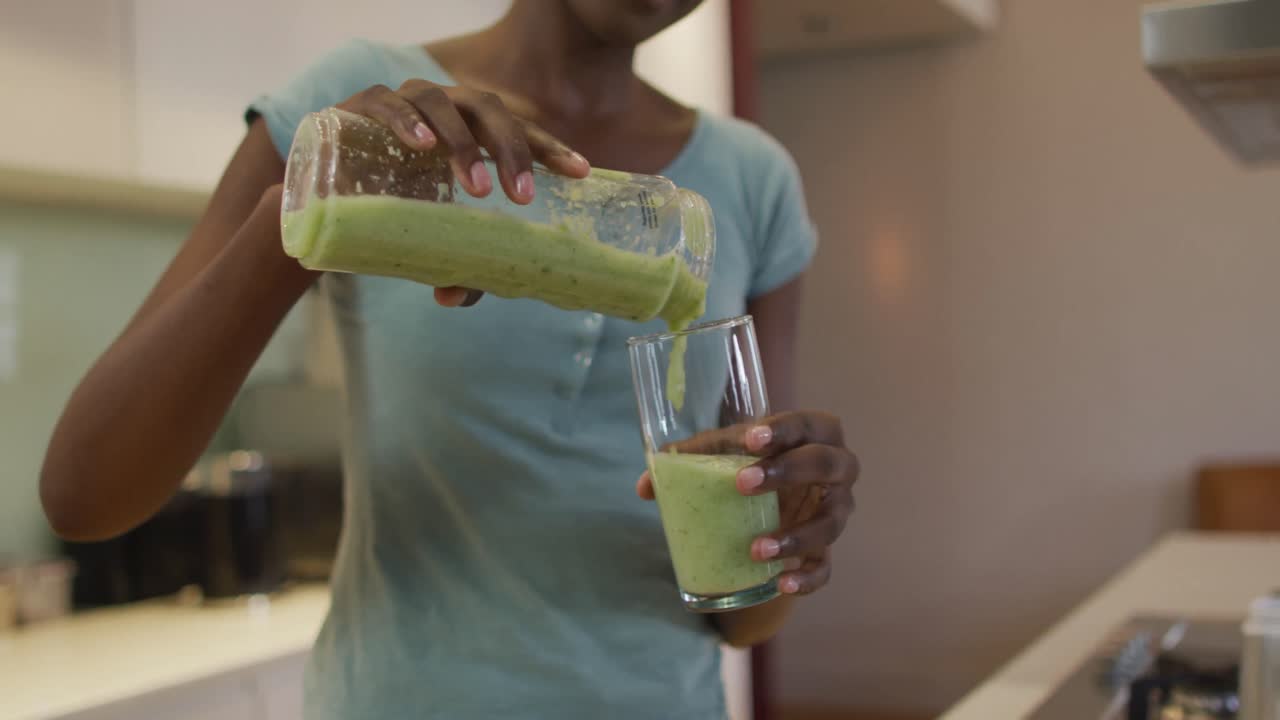 Mani di una donna afroamericana attraente che versa un frullato fatto in casa in un bicchiere in cucina.
