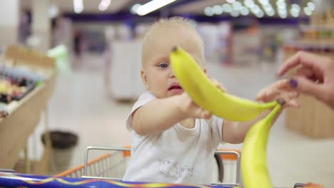 un niño lindo está ayudando a su madre a elegir plátanos en el supermercado mientras está sentado en un carrito de comestibles y sosteniendo plátanos