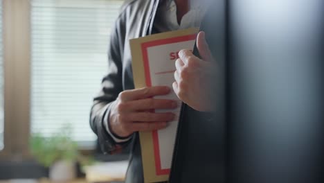 man stealing and hiding top secret file folder in leather jacket inside office. mysterious government agent or spy holding envelope with classified documents and information. taking confidential file