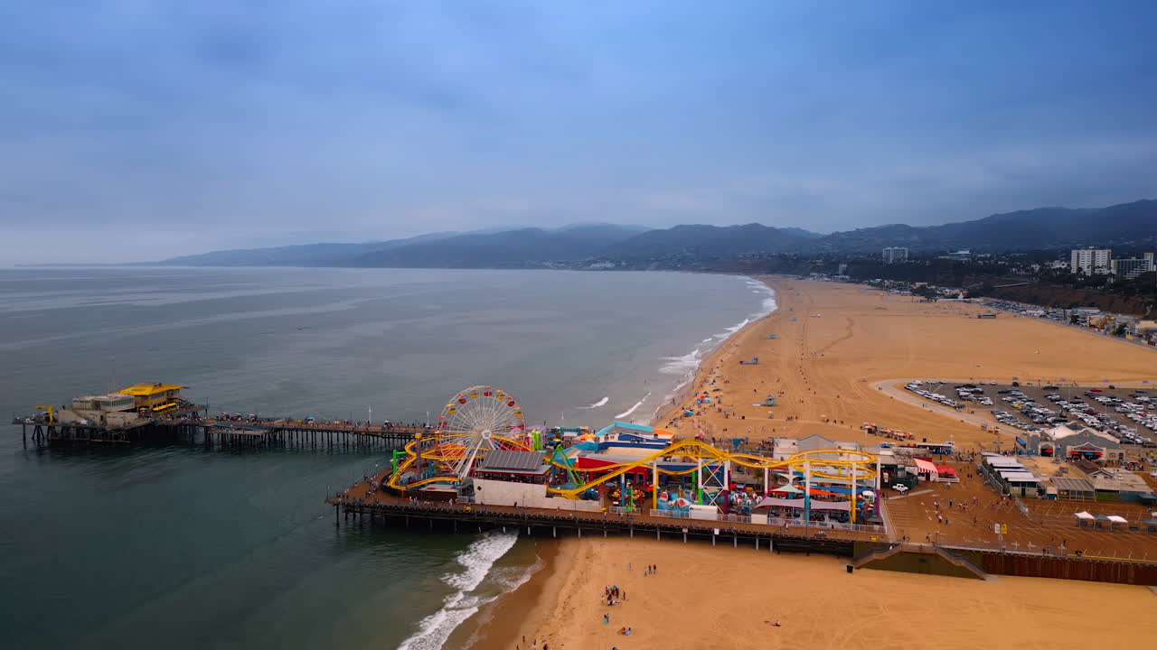 Los Angeles, USA, 29 August 2025: Pacific Park on the Santa Monica Pier from drone footage. Sandy Beach of LA, USA on cloudy day at backdrop