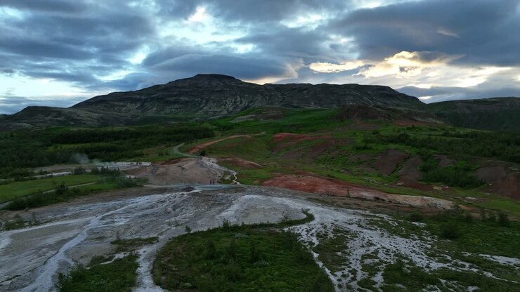 Aerial view of Geysir geothermal area in Iceland, showcasing steaming terrain, vibrant red hills, and a striking mountain backdrop at sunset.
