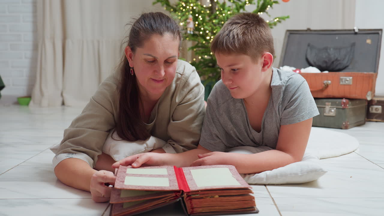 close view boy and mother lying on white floor together looking through open photo book near decorated christmas tree warm cozy atmosphere sharing memories holiday spirit bonding peaceful moment