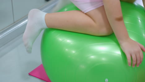 close-up of little child in pink attempting to balance on green stability ball but losing control, child hands press into the ball for support while struggling to maintain stability in an indoor gym