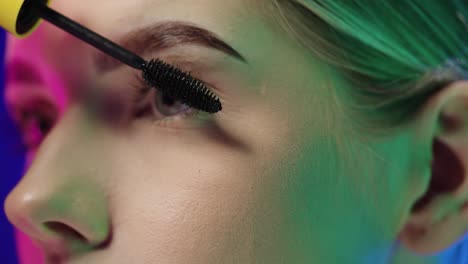 close up of the eyes of a young pretty blonde woman while she is fixing her eyelashes with a mascara before going out or partying in the club in front of blue background in slow motion