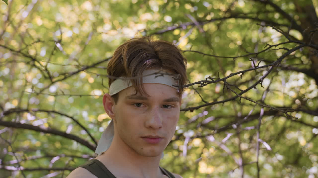 young man standing among slender bare branches in sunlit forest, headband tied around brow, focused gaze into distance, dappled light filtering through green canopy, serene adventurous mood