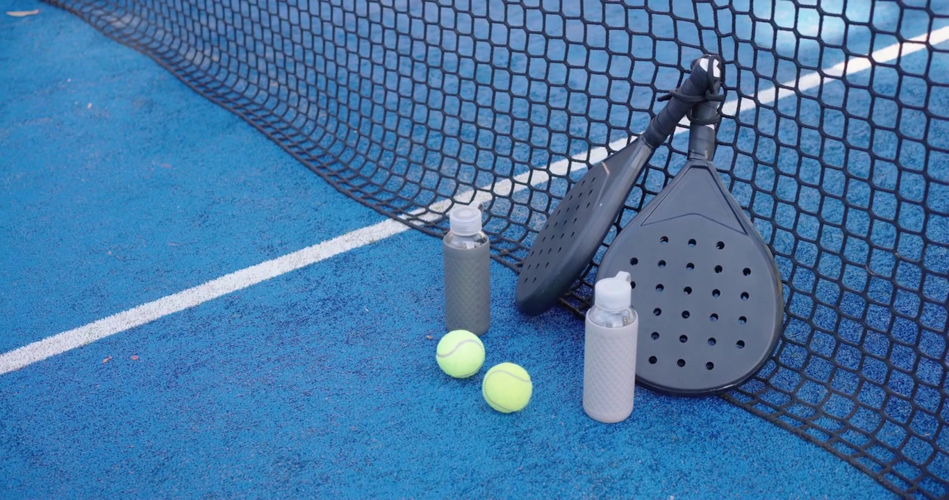 Padel-Schläger, Tennisbälle und Flaschen stehen am Netz, während die Kamera schwenkt und die Ausrüstung zeigt.