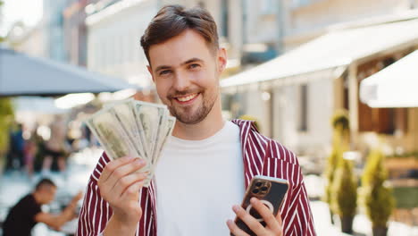 happy bearded man counting money dollar cash use smartphone calculator app in urban city street