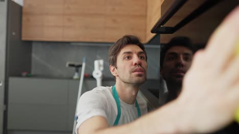 un hombre moreno confiado en una camiseta blanca y un delantal azul es un limpiador lavando una superficie brillante con un trapo amarillo y detergente en la cocina en un apartamento moderno