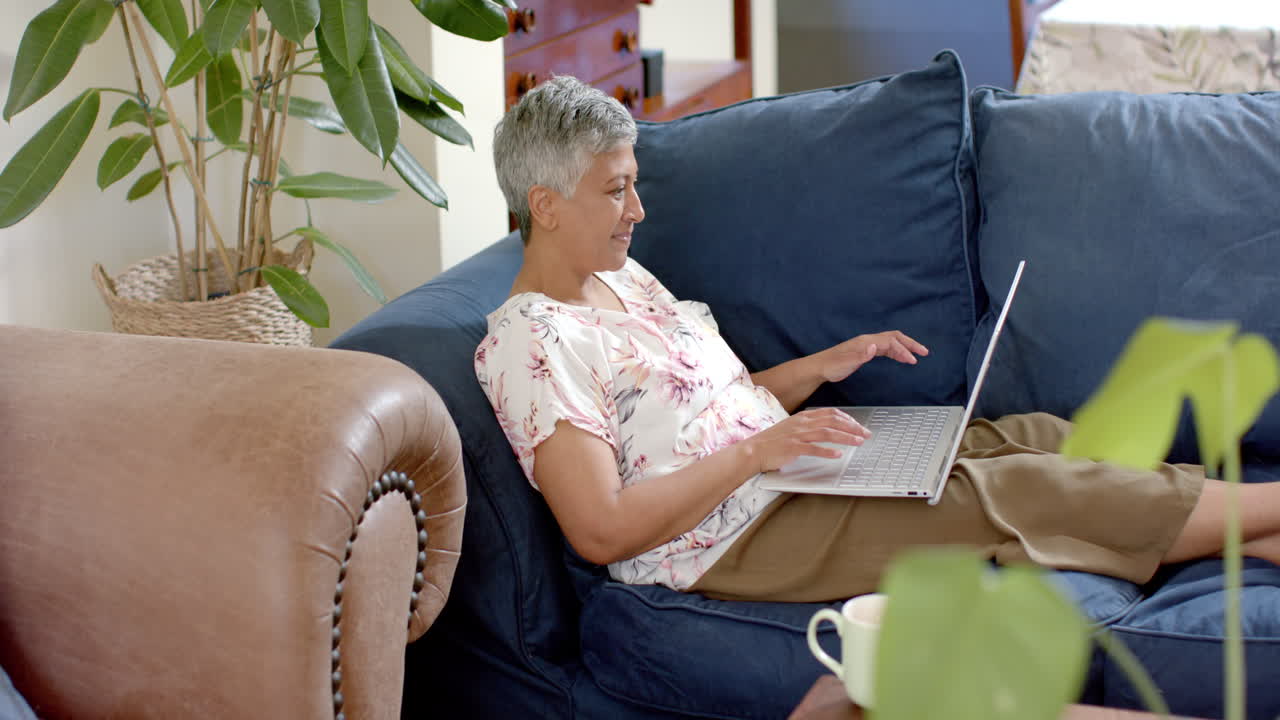 Happy senior biracial woman sitting on couch and using laptop at home, slow motion