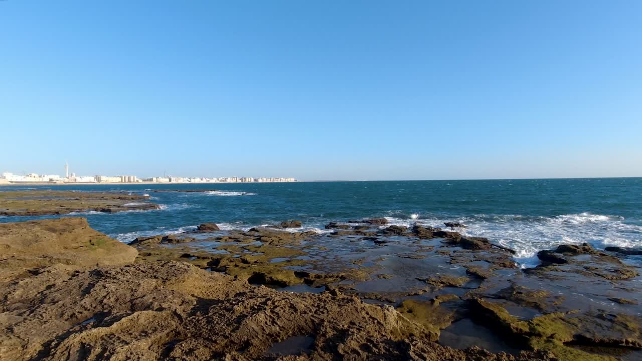 olas rompiendo en el arrecife de piedra con el océano y la ciudad a distancia