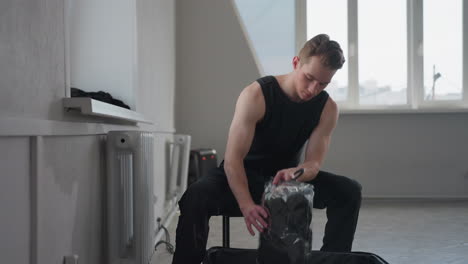 boy in black sleeveless top sits on stool in bright empty room with high window, opens suitcase on floor, retrieves folded garment in plastic sleeve and places it on window ledge before standing