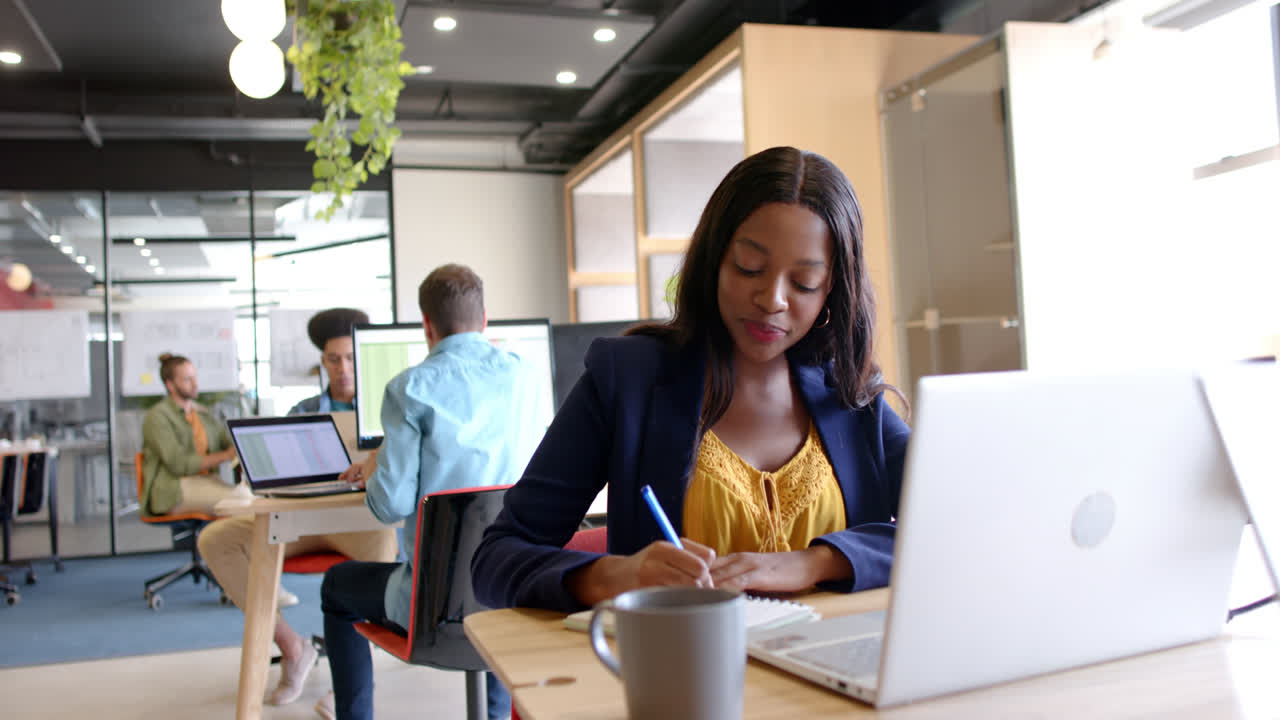 Happy african american casual businesswoman using laptop and writing in casual office, slow motion