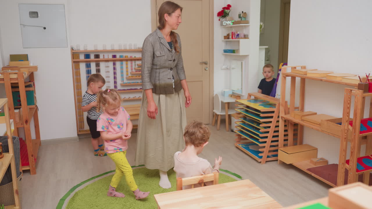 Instructor gathers kindergarten children during classroom activity, encouraging participation and focus while other kids engage in play based learning surrounded by colourful shelves