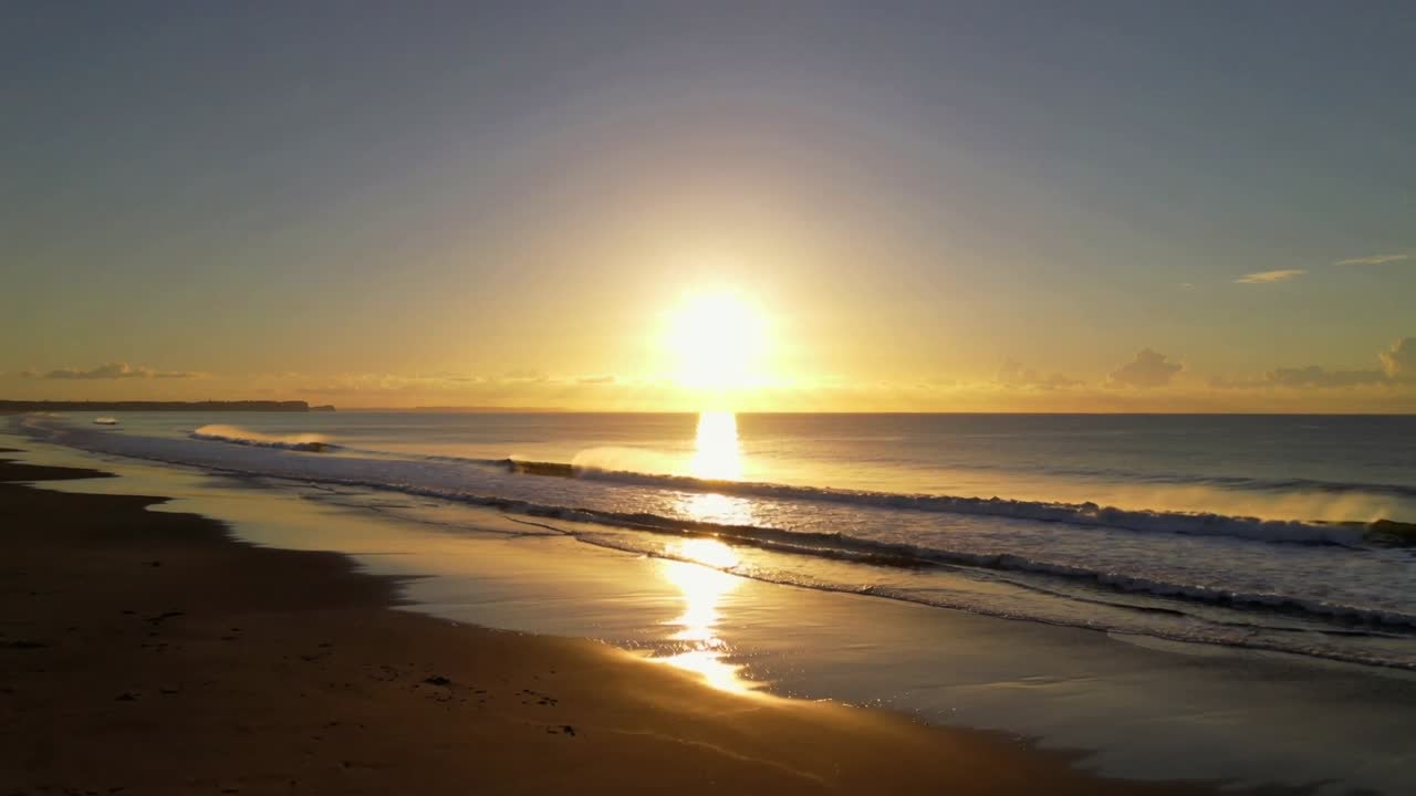 Sol a nascer perto do horizonte projetando um reflexo vertical brilhante na areia molhada da costa, com ondas a quebrar.