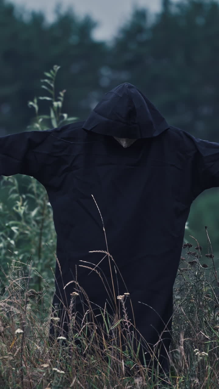 Portrait of ghost with outstretched arms in nature. Scary figure of death in black robe in the field. Horror face of a devil in hood. Vertical video