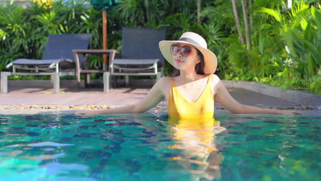 Bonita-Mujer-Asiática-Con-Gafas-De-Sol-Y-Sombrero-De-Paja-De-Pie-En-La-Piscina-Con-Traje-De-Baño-Amarillo-Disfruta-Del-Sol-Mientras-Está-De-Vacaciones