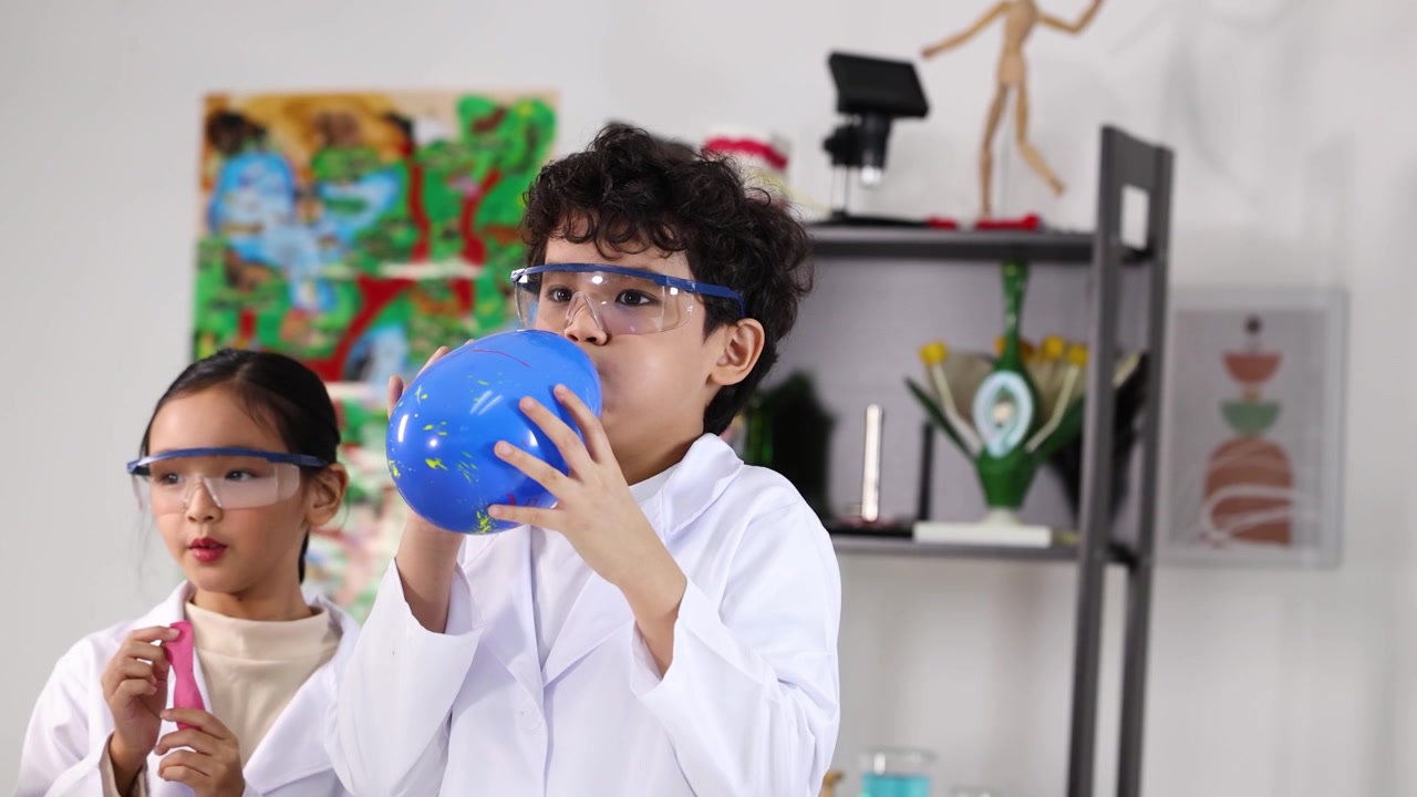 Asian student wearing safety goggles and lab coat blows blue balloon in classroom science lab.
