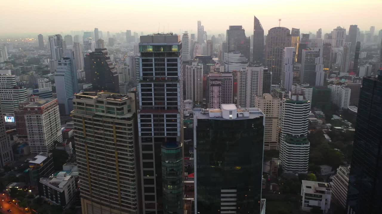 Epic drone flying sideways alongside skyline and skyscrapers of Sukhumvit district during colorful warm sunrise in Bangkok, Thailand.