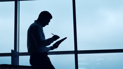 side view of a freelancer sitting on the desk. man in glasses uses stylus pencil working on i-pad. low angle view. wide window at backdrop.