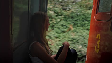 a woman sits at the open door of a moving train on the iconic ella to kandy route in sri lanka, calmly enjoying the stunning landscape of tea plantations, hills, and tropical forest.