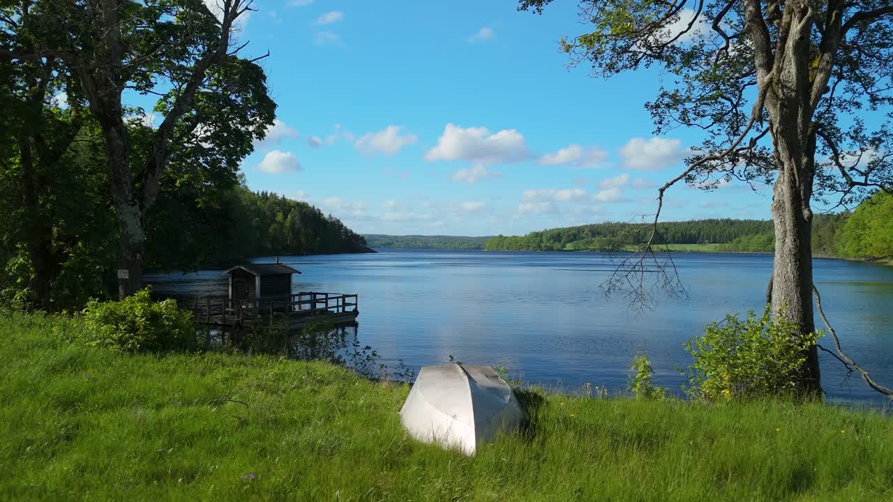 Långsam drönaråkning in mot träbrygga och liten båt på gräsbevuxen strandlinje vid Teåkersjön i lantliga Dalskog Mellerud Sverige under en sommardag