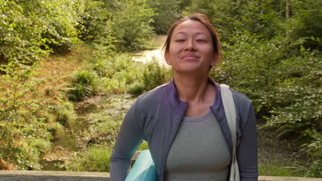 mujer con ropa deportiva llevando una alfombra de yoga de pie en un puente en el bosque mirando a su alrededor disfrutando de la paz y la belleza de la naturaleza 5