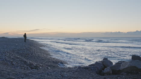 man walks along stoney shore in early morning as waves wash up onshore