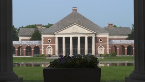 a beautiful building and spring sit in the sunlight beyond two columns with a stone flower pot in the foreground as the camera pulls back