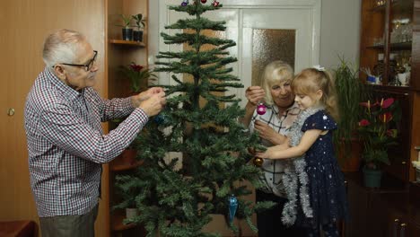 Children-girl-with-elderly-couple-grandparents-decorating-artificial-Christmas-pine-tree-at-home