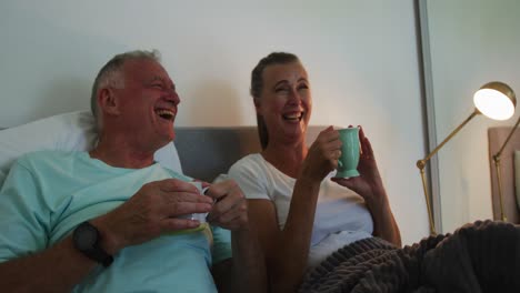 pareja de ancianos caucásicos riendo mientras beben una taza de café en la cama en casa