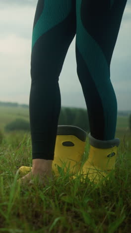 mujer en traje verde y leggings levanta su pie desnudo para quitar la bota amarilla mientras está de pie en un vasto campo de hierba con árboles en la distancia