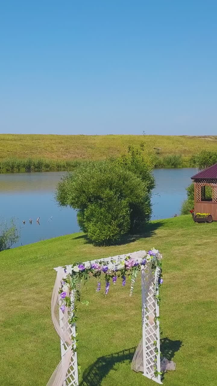 stylish wedding venue with decorated arch on lush green meadow near large tranquil pond on sunny day aerial view
