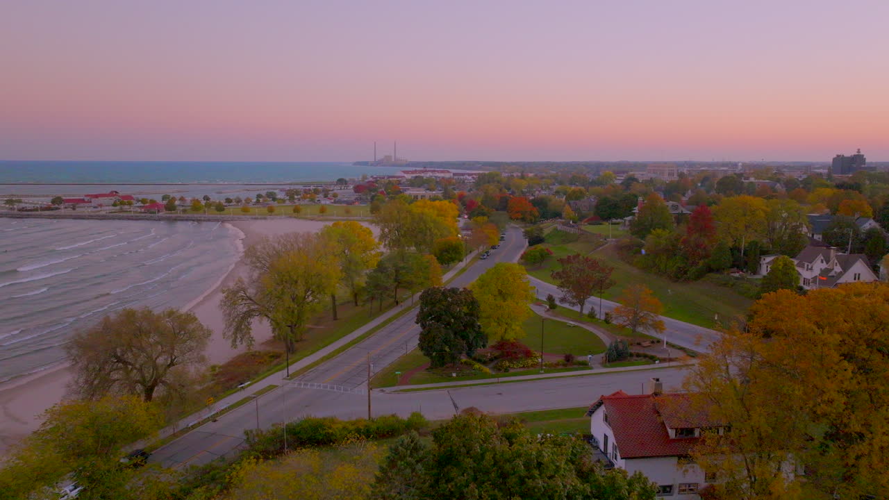 Drone aerial pushing over a charming Sheboygan, Wisconsin neighborhood with colorful trees and an American flag waving beside Lake Michigan toward a pink and purple ombré sunset sky