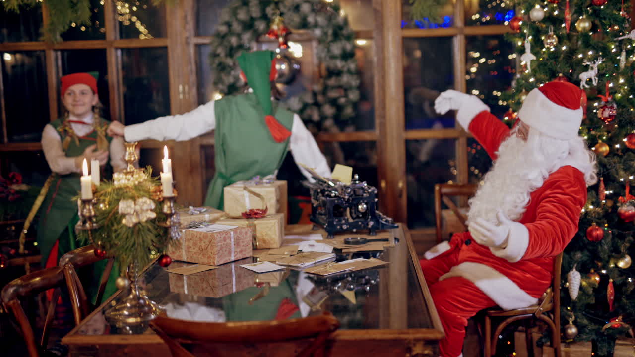 Santa sitting at the table and two elves dance together. Happy elves in fairy costumes dancing while Santa Claus is watching at them on christmas background indoors.
