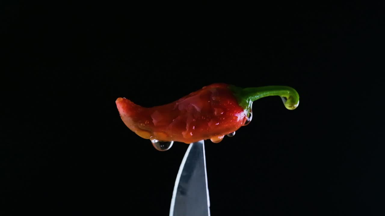 un pimiento de chile rojo encaramado en el extremo de un cuchillo afilado con agua goteando sobre él en cámara lenta