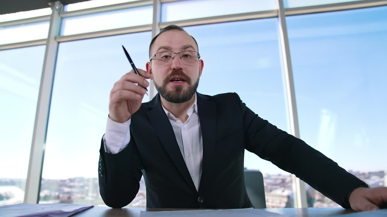 Portrait of a businessman sitting in office. Bearded entrepreneur in suit and glasses talking about business on window backdrop.