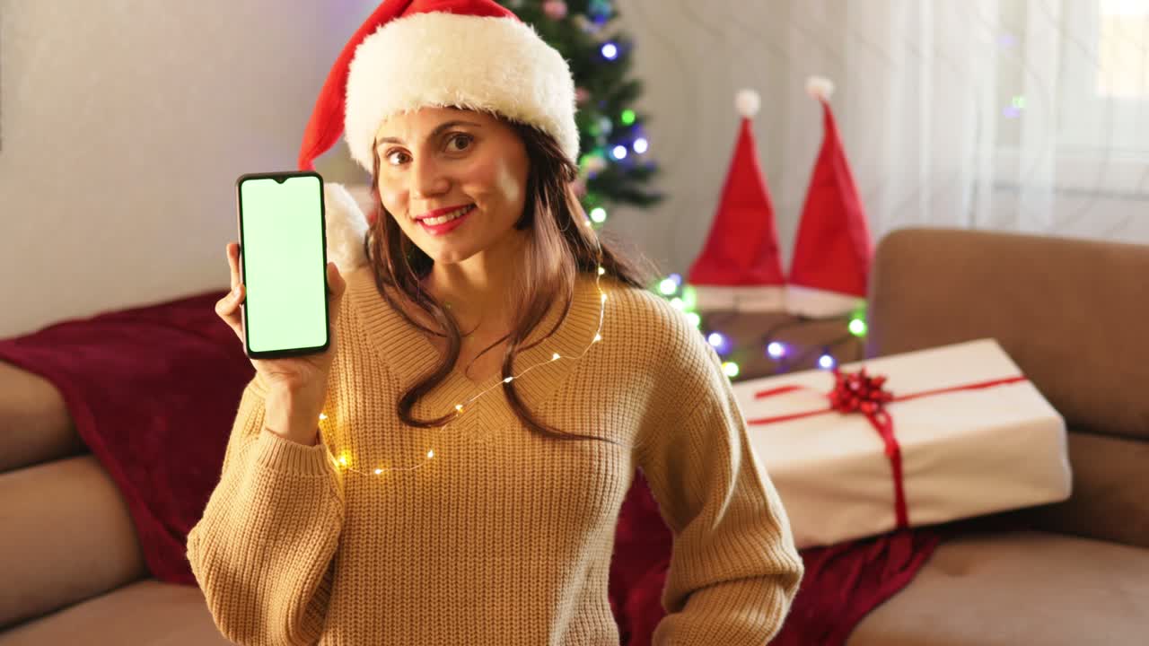 feliz mujer sonriente en el sombrero de santa claus sosteniendo un teléfono móvil con un fondo verde sentada en el sofá en el interior acogedor de navidad. celda de pantalla verde
