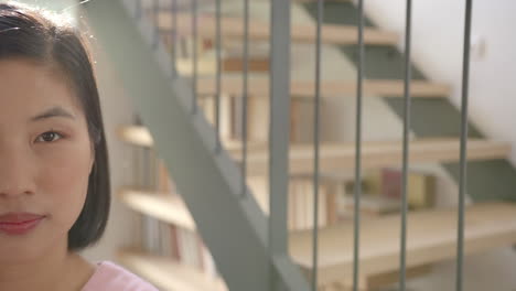 smiling asian woman standing near staircase with bookshelves in modern home