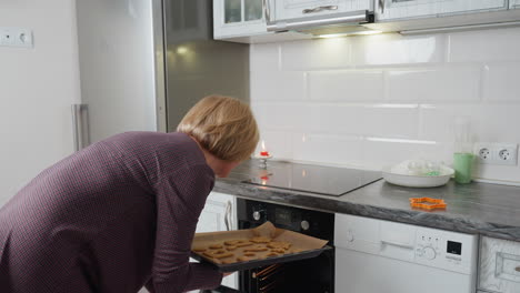 mujer abriendo el horno y preparando para colocar las galletas en la bandeja de horneado, con equipos de cocina como rodillo, cortador de galletas y vela en la mesa en el fondo