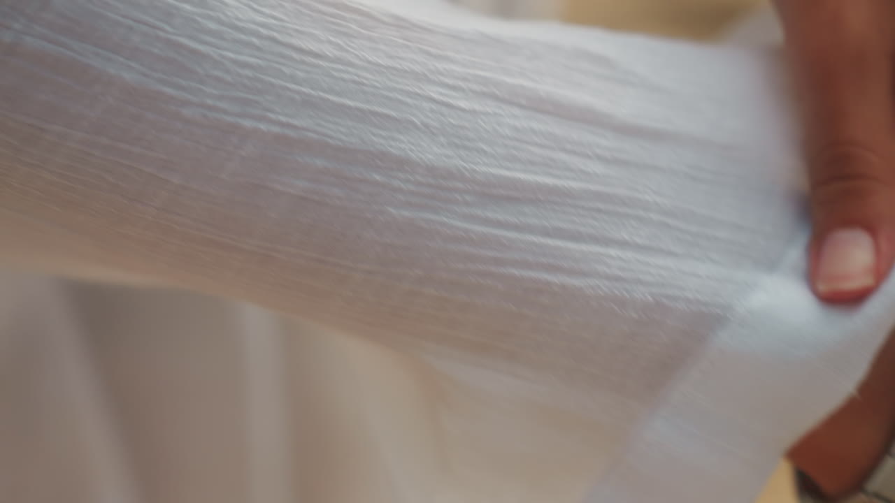 Nahaufnahme von Händen, die am Meer bei Sonnenuntergang einen weißen Hemdärmel hochkrempeln, Finger glätten Leinen, befestigen Knopf und Uhr, während ein Ring warmes Licht einfängt – ein lässiger Styling-Moment für Urlaub oder Fotoshooting.