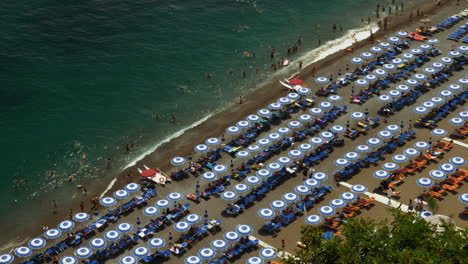 Looking-down-upon-a-summer-beach-scene-with-beautiful-clear-water,-people-playing-in-the-sand,-and-the-beach-covered-in-umbrellas