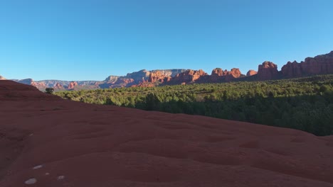 Drone-flys-along-sandstone-ledge-over-forest-to-Submarine-Rock,-Sedona-Arizona