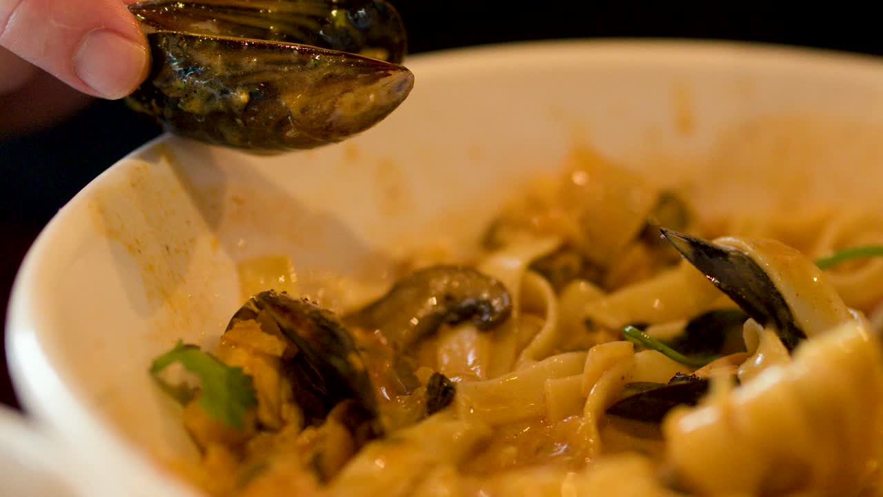 Close-up of hand holding mussel above creamy seafood pasta, warm lighting, shallow depth of field